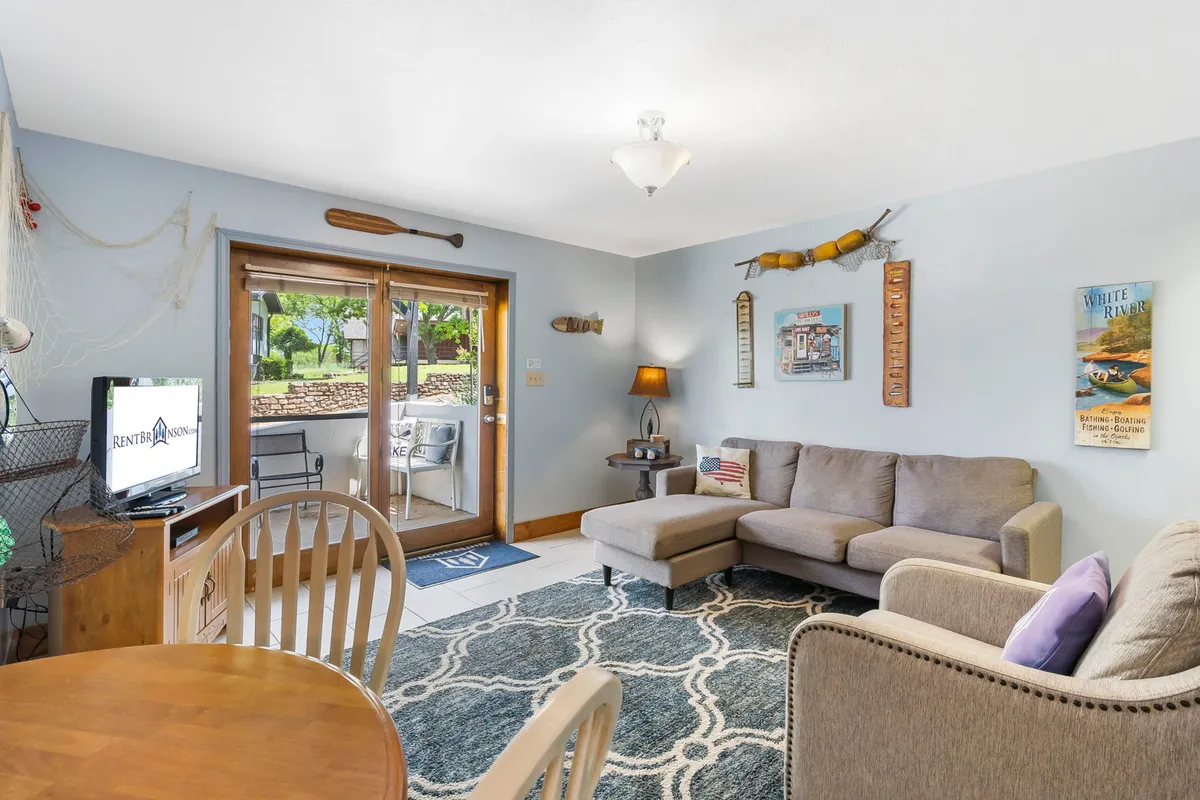 The image captures a warmly lit, ground-level living space with a cozy sofa, a modern dining table set for gathering, a television for entertainment, and a large sliding door that offers a view toward the lake and sunset. Artistic wall décor adds a welcoming touch, while the layout invites relaxation and quality time together.