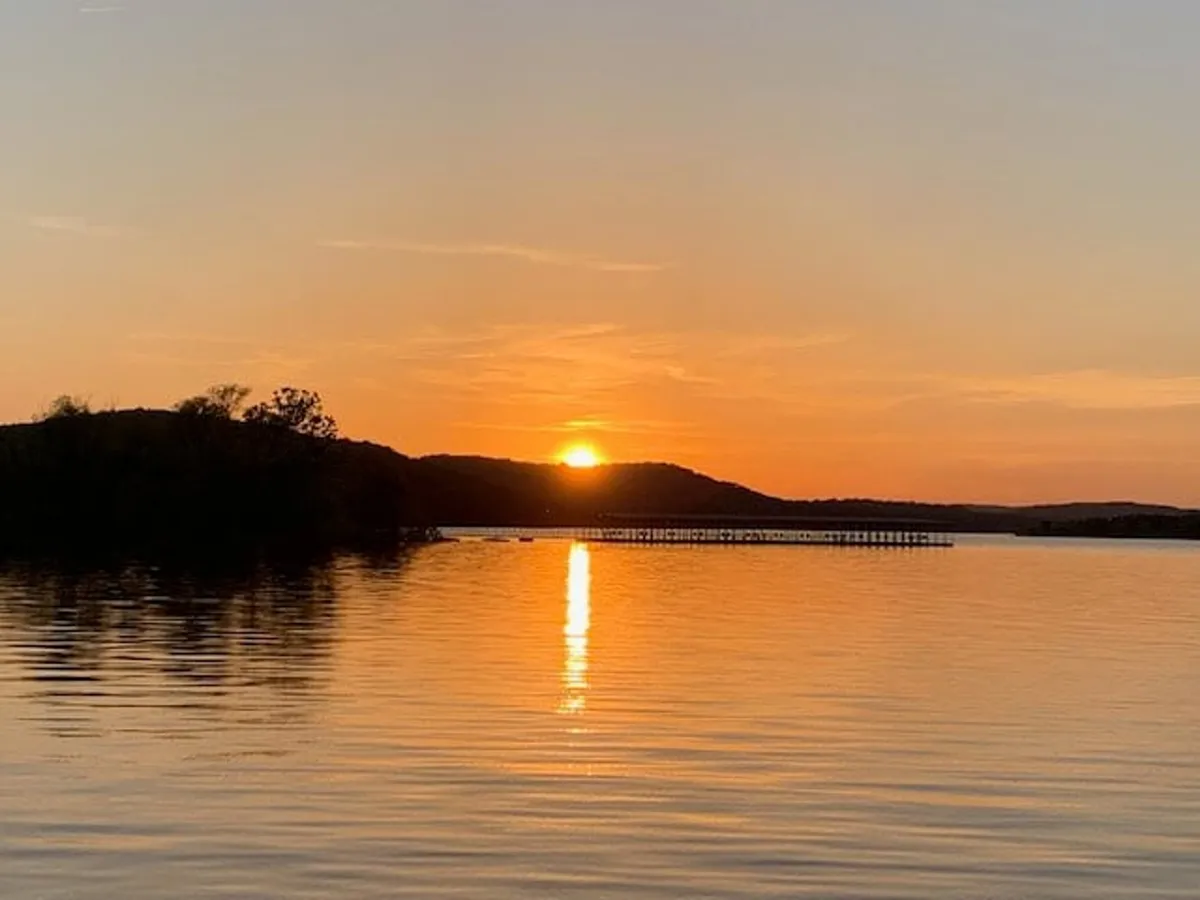 A tranquil sunset scene over Table Rock Lake, with calm waters gently reflecting the sky's warm hues. In the distance, a pier extends into the lake, and the silhouette of rolling hills creates a peaceful backdrop, emphasizing the lakeside setting.