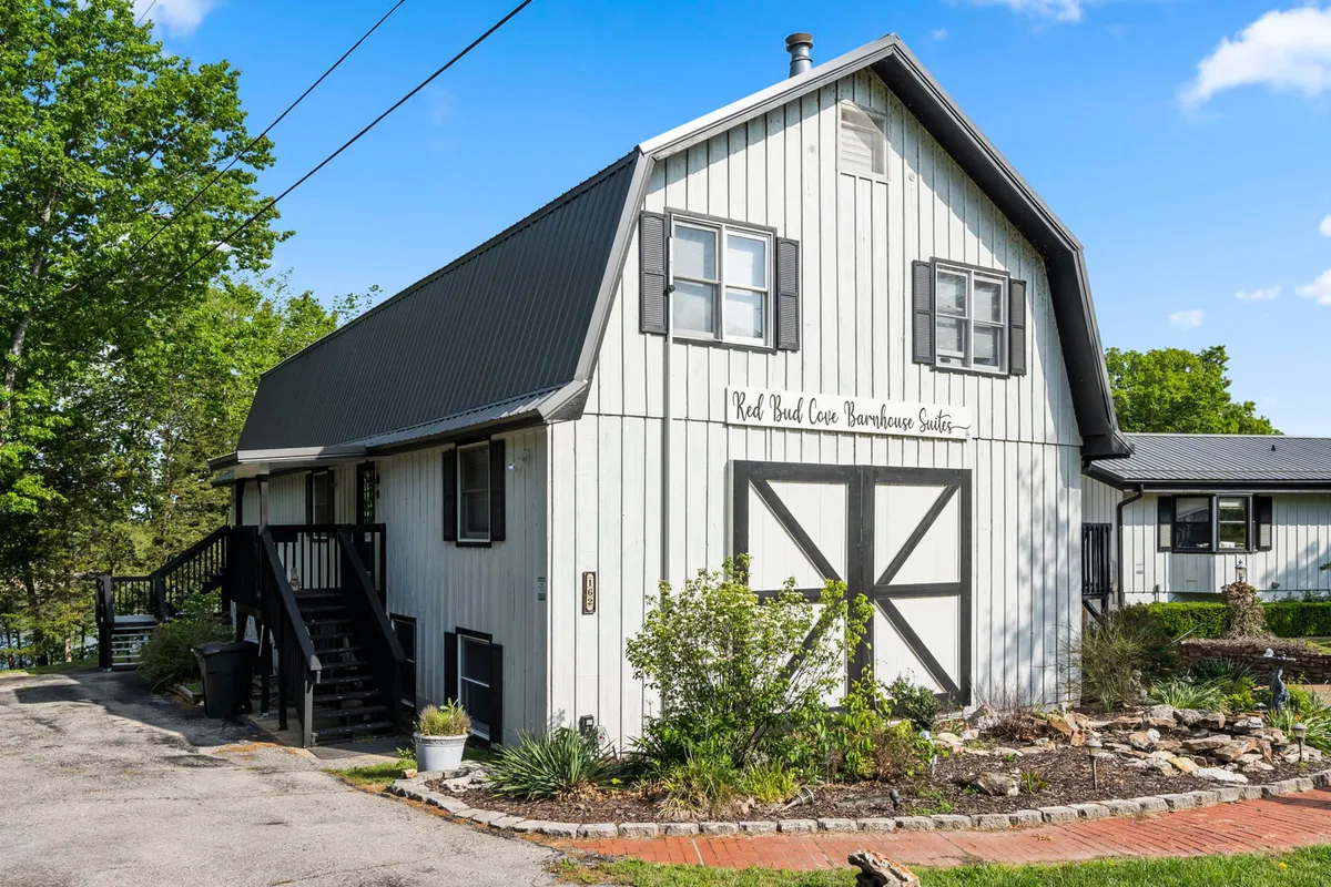 A charming garden scene with barn-style architecture and clear signage for 'Red Bud Cove Barnhouse Suites,' framed by landscaping that hints at access to nature trails and lakeside areas.