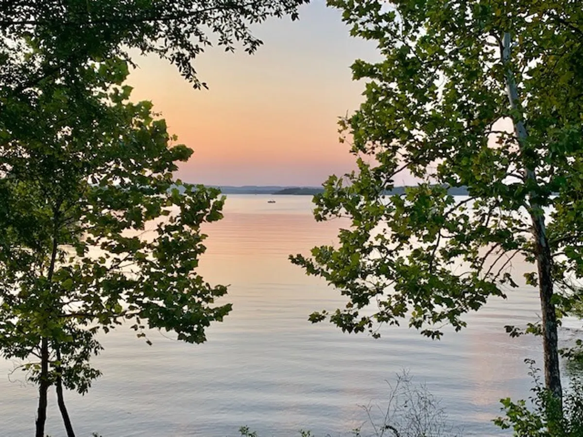 A tranquil view of a calm lake framed by trees with a stunning sunset sky overhead. The natural setting evokes relaxation and recreation, inspiring thoughts of swimming, fishing, or simply gathering to enjoy the water’s edge and the shared hot tub nearby.