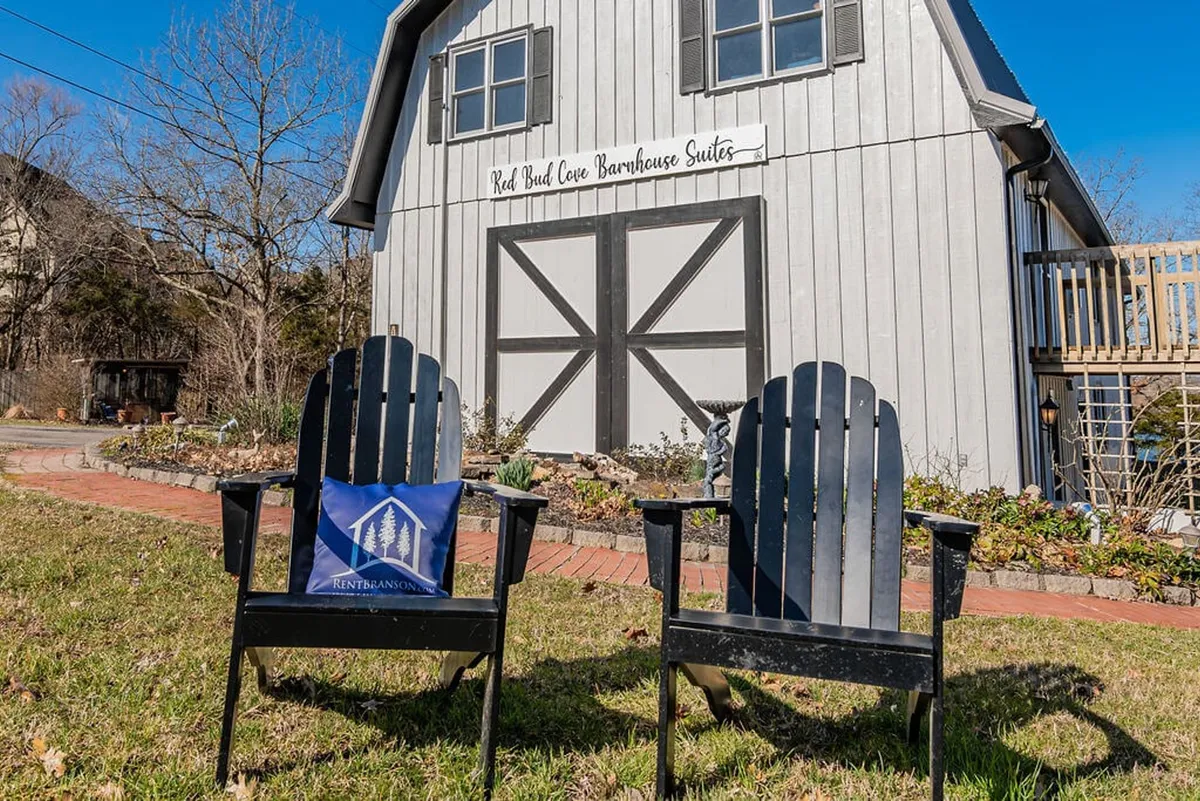 A welcoming barn-style building sits at the water’s edge, with wooden chairs and a decorative pillow arranged for comfort. The exterior scene captures the calm atmosphere of the lakefront, perfect for watching the sunset or sharing a quiet moment with a companion.