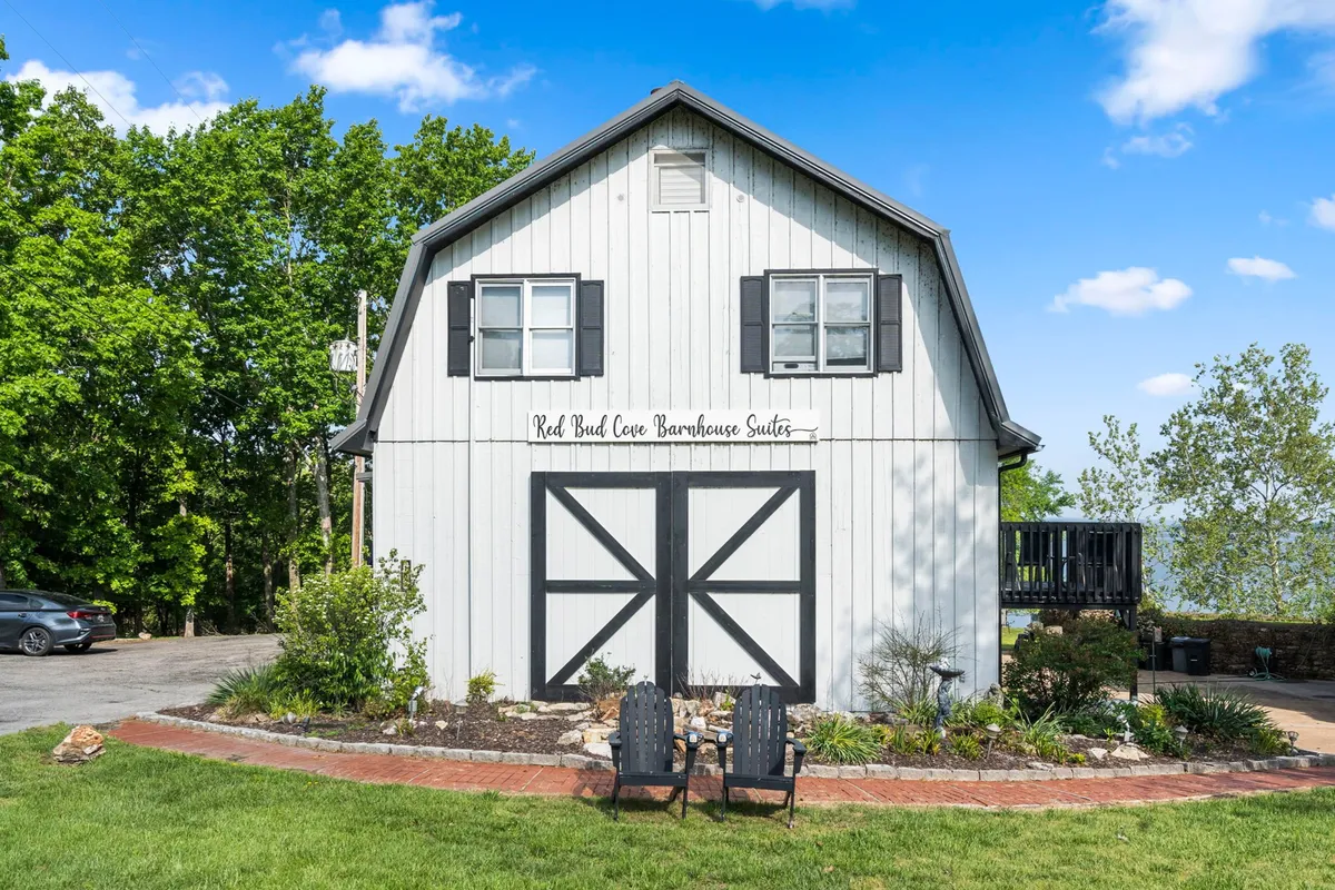 The photo showcases the barnhouse exterior with double doors opening onto a charming seating area surrounded by landscaping. The setting is calm, inviting, and perfect for relaxing outdoors or gathering with others after a day of lakeside activities.