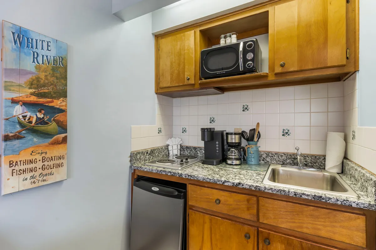 A cozy kitchenette with granite countertops, wooden cabinets, modern kitchen appliances, and vintage wall art. The countertop holds a coffee maker and cooking essentials, and the space is warm and inviting, ideal for preparing meals.