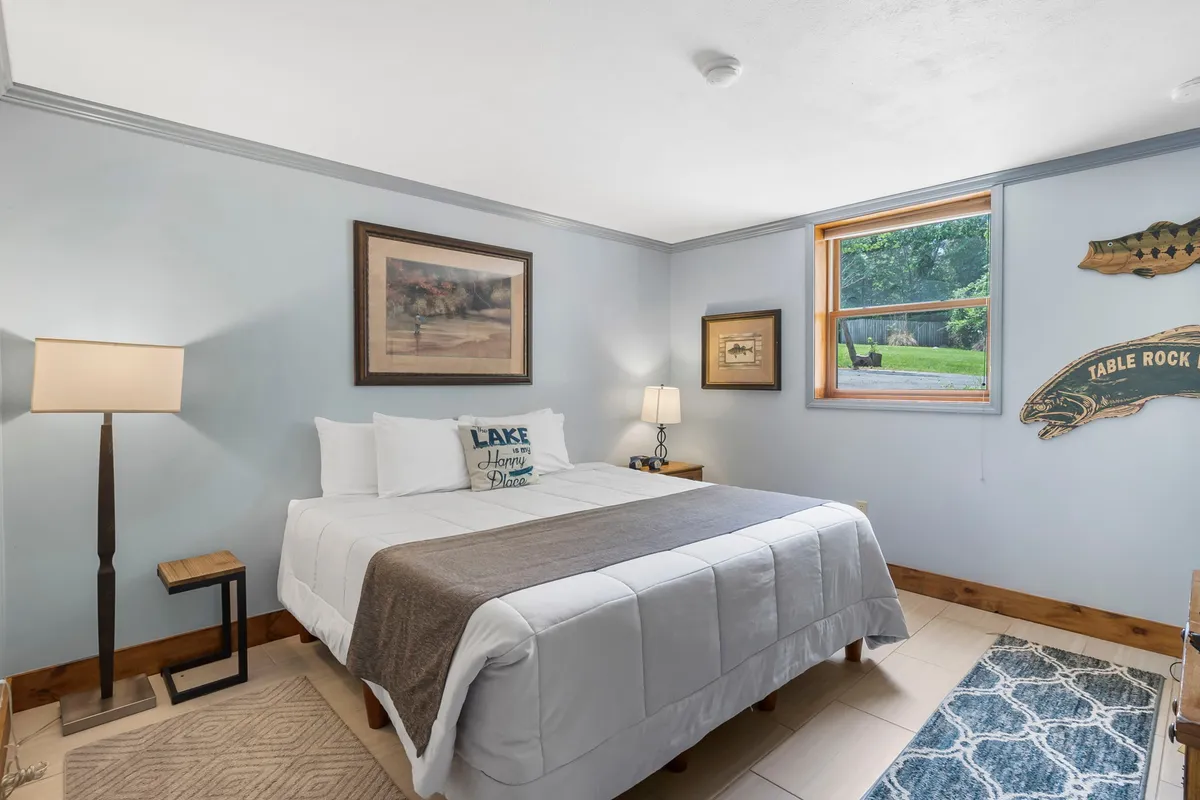A serene bedroom featuring a bed with decorative pillows, framed artwork, a bedside lamp, and a window offering natural light and outdoor views. A whimsical fish plaque adds a charming touch.