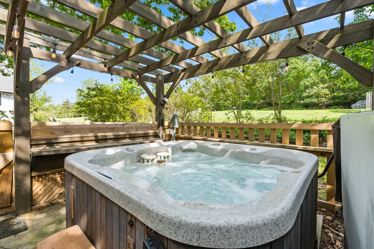 This image features a shared outdoor hot tub set under a wooden pergola, surrounded by lush greenery. People can be seen relaxing in the jacuzzi and socializing, creating a tranquil yet sociable atmosphere. The area is ideal for both daytime relaxation and nighttime stargazing, framed by a well-maintained landscape.
