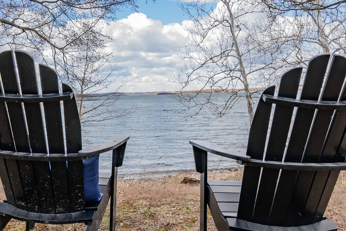 The image shows two black Adirondack chairs set on a patio overlooking a calm lake and expansive sky. The scene exudes tranquility and invites guests to relax, read, or enjoy the view as the sun sets over the water.