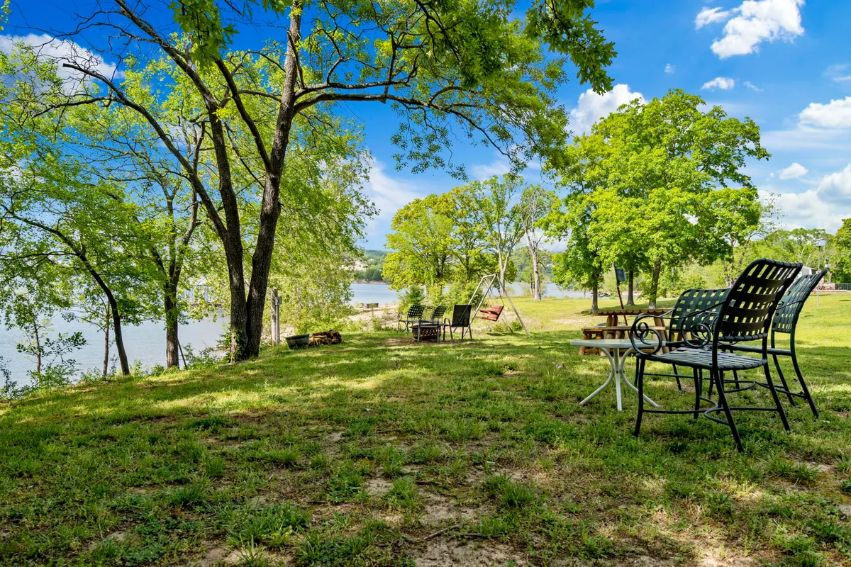A pair of chairs sit beneath leafy trees on the riverbank, overlooking calm water. The scene is peaceful, inviting guests to relax, fish, or picnic with direct access to the water's edge.