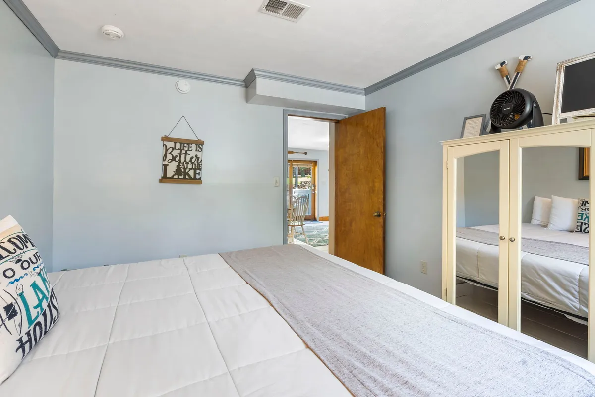 A spacious bedroom with a king bed dressed in crisp white linens and a decorative pillow, with soft morning sunlight filtering in. The room includes a mirror cabinet, wall artwork, and an open door that hints at connected living spaces.
